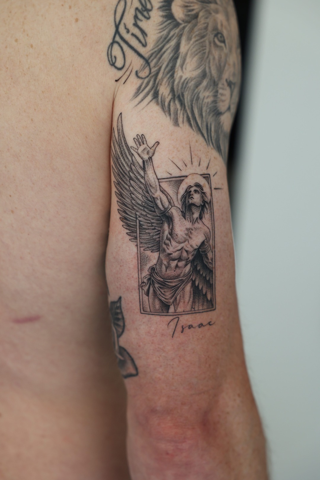 Winged figure reaching towards the light with Isaac lettering, upper arm tattoo, black and grey by Matt Roe, Kensington London