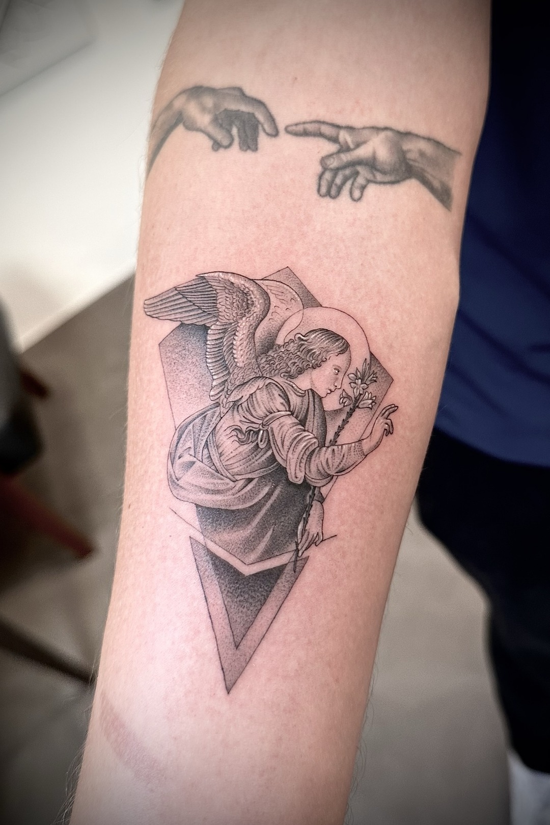 Annunciation angel holding a lily with geometric framing, forearm tattoo, black and grey by Matt Roe, Kensington London