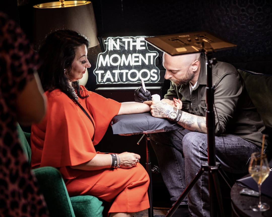 Matt Roe tattooing a guest's hand beneath the neon sign