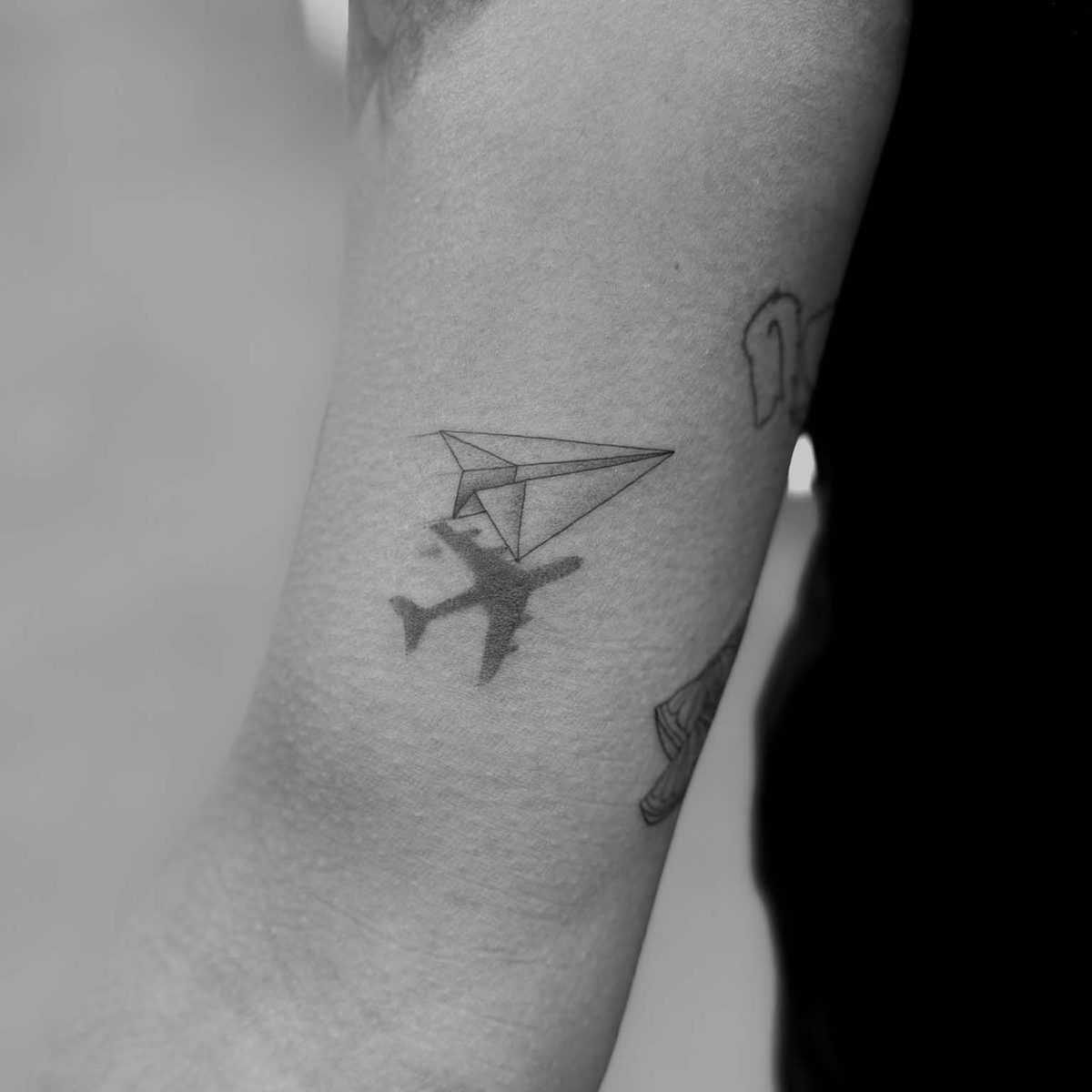 Fine line paper plane with airplane shadow tattoo on bicep by Matt Roe, London